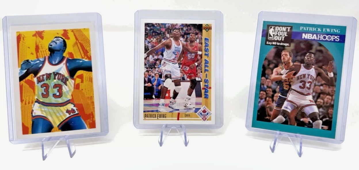 Three Patrick Ewing New York Knicks cards in clear plastic top loaders on acrylic stands against a white backdrop: the yellow-and-orange "New York 33" art card, the white-bordered East All-Star action shot, and the teal NBA Hoops "Don't Foul Out" card